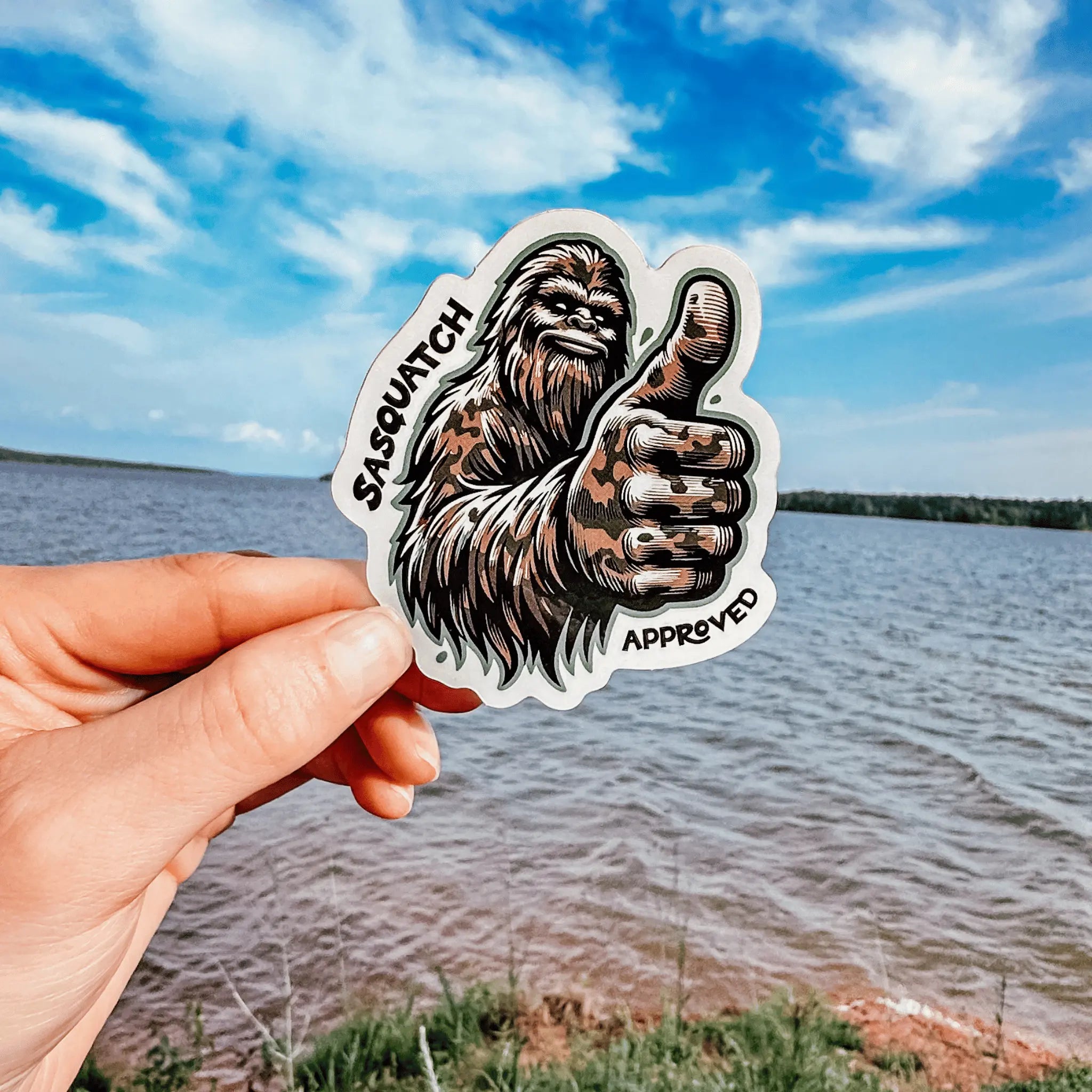 Hand holding the Squatch Approved Sticker outdoors by a lake — rugged waterproof Bigfoot decal showing a thumbs-up, perfect for gear, bottles, and adventure life.